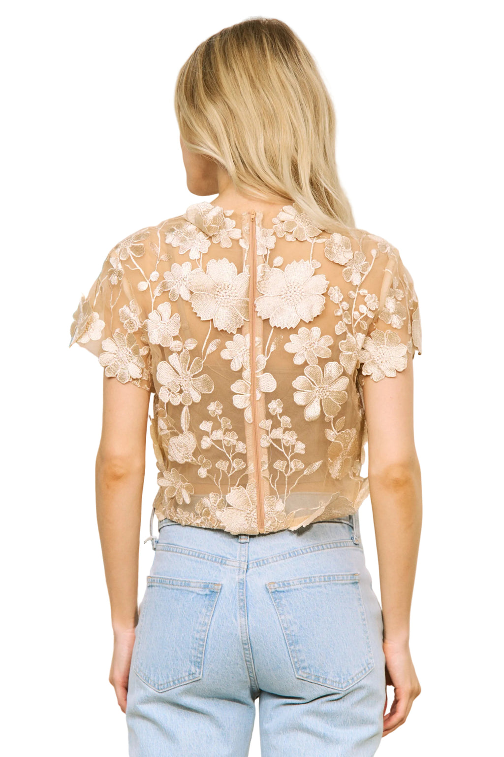 Woman wearing a sheer top with floral embroidery and light blue jeans indoors.